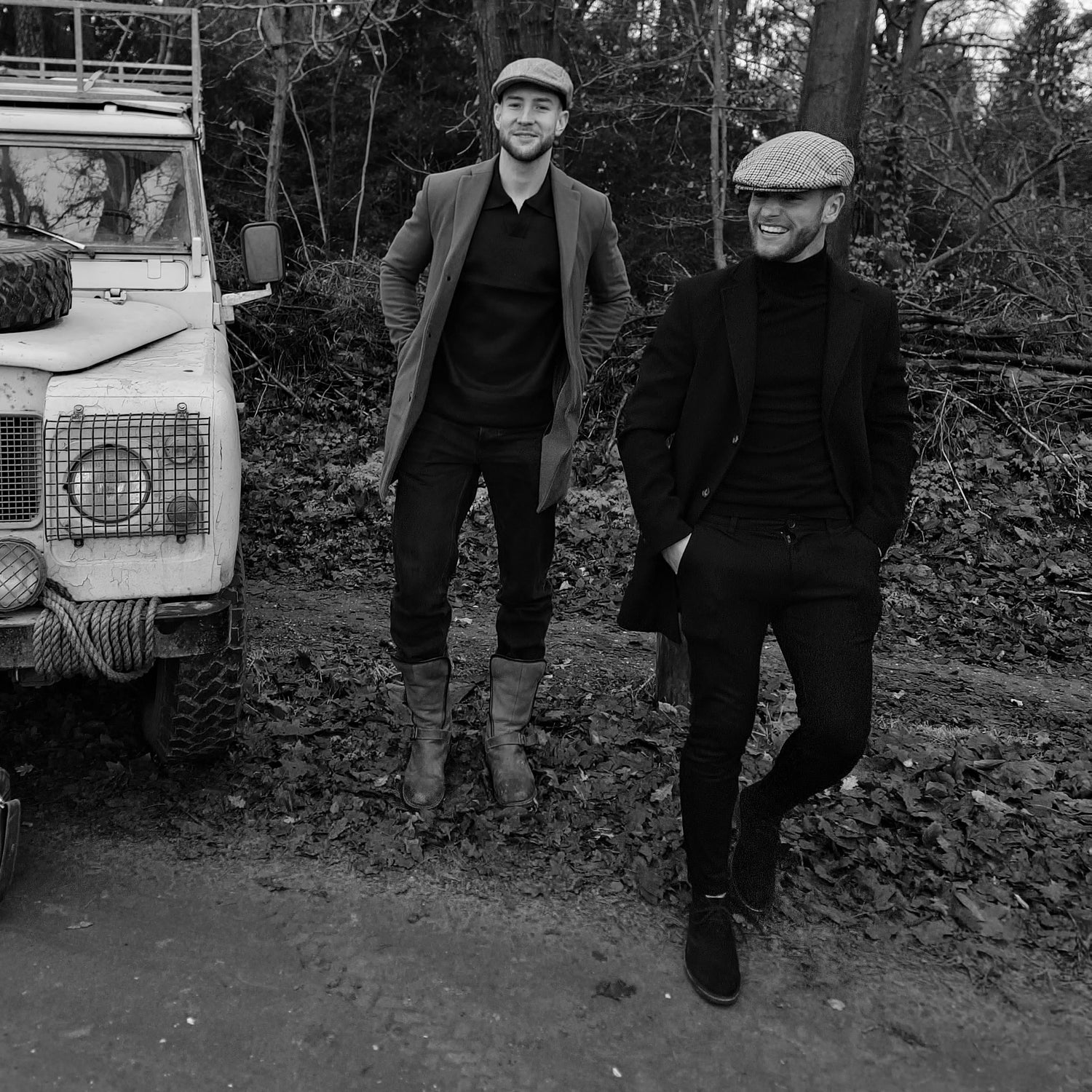 Tweetal mannelijke modellen die lachend weglopen bij een robuuste SUV. De foto is deel van een fotoshoot van het merk ATSORT, in de oud-engelse stijl. Beide modellen dragen een engelse flatcap flat cap.